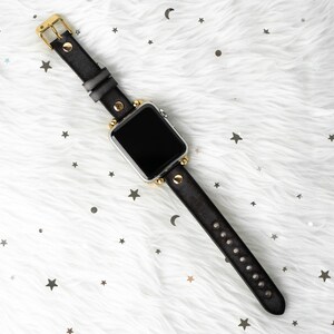 Puede incluir: Una correa de reloj de cuero negro con detalles dorados y tachuelas. La esfera del reloj es negra y la correa est&aacute; sobre un fondo blanco peludo.