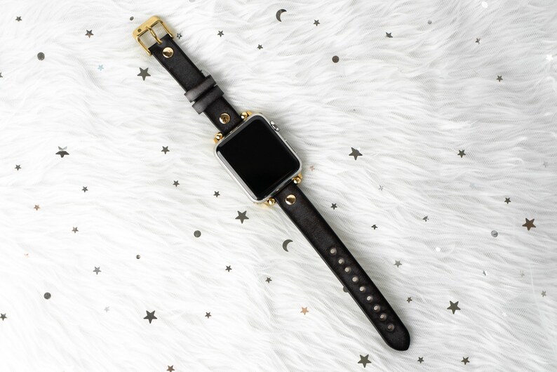 Puede incluir: Una correa de reloj de cuero negro con detalles dorados y tachuelas. La esfera del reloj es negra y la correa est&aacute; sobre un fondo blanco peludo.