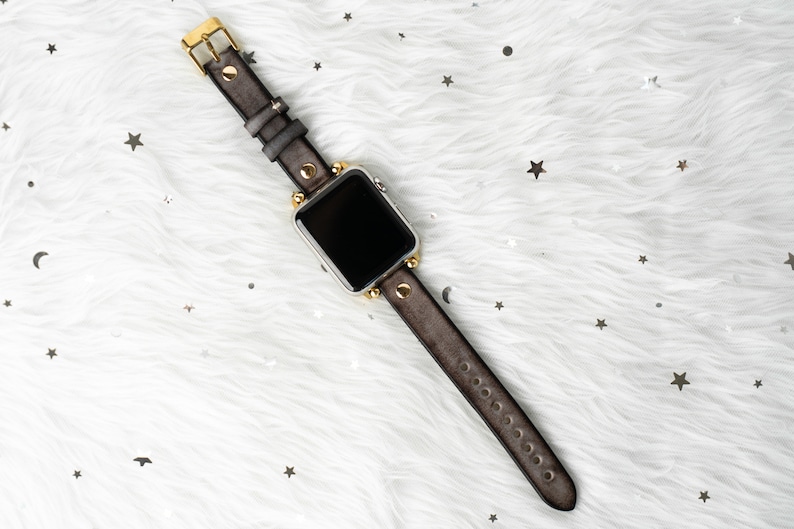 Puede incluir: Una correa de reloj de cuero gris con detalles dorados y una esfera de reloj inteligente plateada.