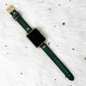 Puede incluir: Una correa de reloj de cuero verde con detalles dorados y una esfera de reloj inteligente plateada.