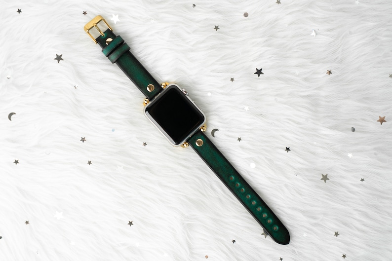 Puede incluir: Una correa de reloj de cuero verde con detalles dorados y una esfera de reloj inteligente plateada.