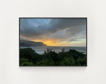 May include: A scenic landscape photograph of a coastline with a mountain range in the background. The sky is a mix of dark gray clouds and a bright yellow sunset. The ocean is a calm blue with white waves crashing on the shore. Lush green trees and foliage are in the foreground.