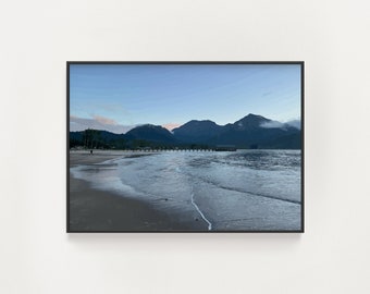 Puede incluir: Una vista panorámica de una playa con agua azul tranquila que baña la orilla. La playa es arenosa y hay montañas en la distancia. El cielo es azul claro con algunas nubes.