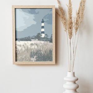 May include: A framed print of a lighthouse on a beach with a cloudy sky. The lighthouse is black and white with a white top. The beach is covered in tall grass. The print is in a light wood frame.
