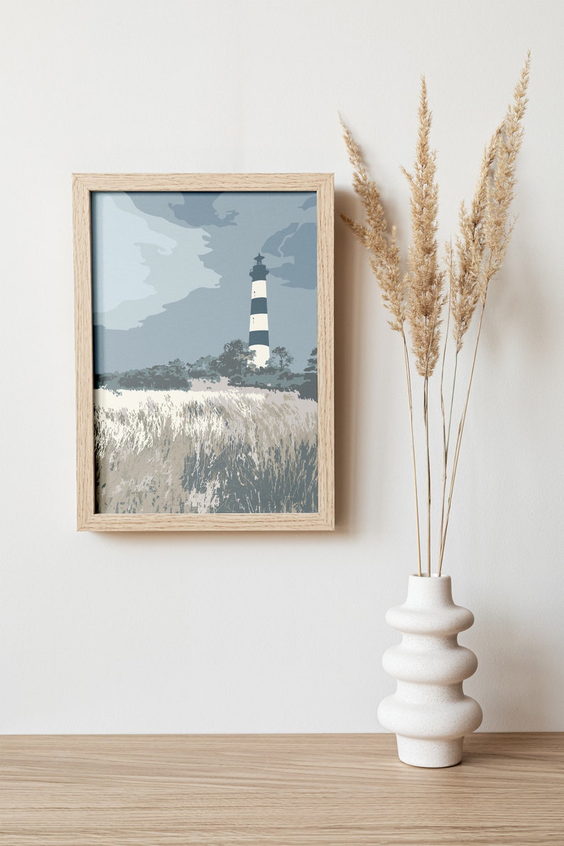 May include: A framed print of a lighthouse on a beach with a cloudy sky. The lighthouse is black and white with a white top. The beach is covered in tall grass. The print is in a light wood frame.