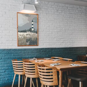 May include: A wooden dining table with six chairs, set against a white brick wall with a framed print of a lighthouse. The table has a light wood finish and the chairs have a natural wood finish.