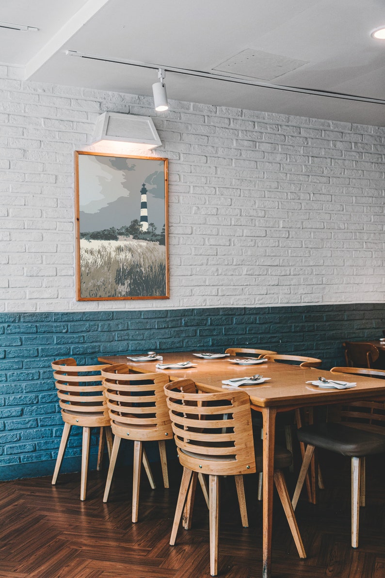May include: A wooden dining table with six chairs, set against a white brick wall with a framed print of a lighthouse. The table has a light wood finish and the chairs have a natural wood finish.