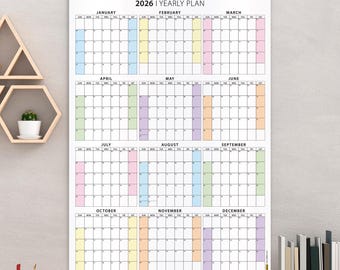 May include: A 2026 yearly plan calendar with a white background is displayed on a wall. The calendar is divided into months with color-coded sections. A wooden desk with office supplies and a lamp is in the foreground.