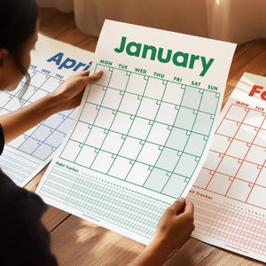 May include: A person holding a large white calendar sheet labelled "January" with green text and boxes. The calendar includes a habit tracker at the bottom. Other calendar sheets for "April" and "February" are visible.