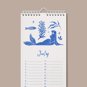 May include: A spiral-bound calendar with a blue mermaid illustration and the word "July". The calendar features a white background with blue illustrations of a fish, seaweed, and stars. The calendar has a monthly layout.