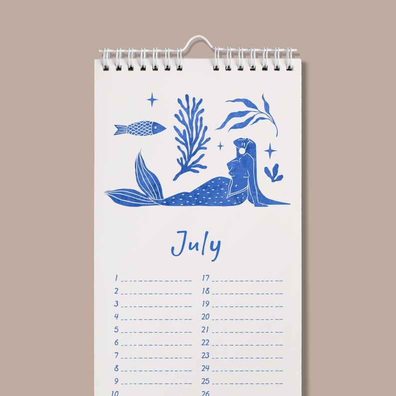 May include: A spiral-bound calendar with a blue mermaid illustration and the word "July". The calendar features a white background with blue illustrations of a fish, seaweed, and stars. The calendar has a monthly layout.