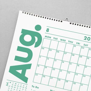 May include: A white wall calendar with a teal green August header. The calendar grid features dates, days of the week, and space for notes. The calendar is spiral-bound and includes a metal hanger.