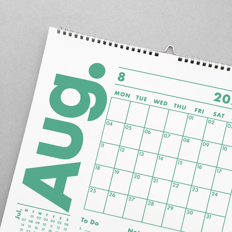 May include: A white wall calendar with a teal green August header. The calendar grid features dates, days of the week, and space for notes. The calendar is spiral-bound and includes a metal hanger.