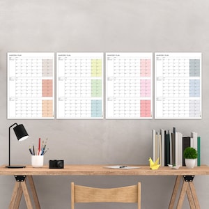 Könnte beinhalten: Vier Quartals-Planer-Poster an einer grauen Wand. Jedes Poster hat ein anderes Pastellfarbschema: Pfirsich, Gelb, Rosa und Blau. Die Poster hängen über einem Holztisch mit einer Lampe, Bleistiften, einer Kamera und einem Laptop.