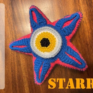 May include: A crocheted plush star with a large eye in the center. The eye is yellow, white, and black. The star is pink and blue.