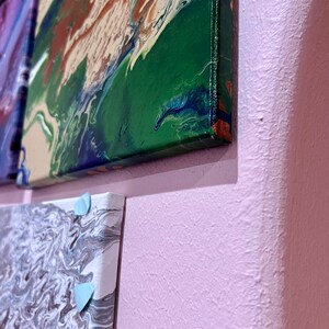 以下が含まれることがあります： 緑、青、茶色の抽象画のクローズアップ。キャンバスに描かれた絵は、渦巻き状で流動的なデザインです。キャンバスは壁に取り付けられています。