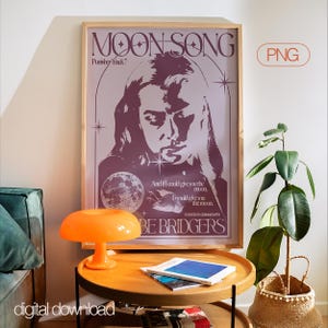 May include: A framed poster with the text "MOON SONG" and an illustration of a person's face, a moon, and the words "BE BRIDGERS". An orange mushroom lamp sits on a wooden table with books. A plant in a woven basket is next to the table.