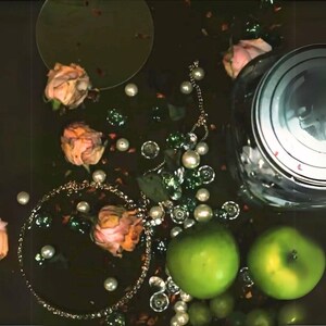 Peut inclure: Une nature morte avec un bocal en verre avec un couvercle en forme de cr&acirc;ne, deux pommes vertes, un collier en cha&icirc;ne dor&eacute;e avec des perles et plusieurs roses roses sur un fond sombre.