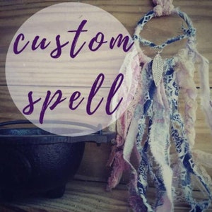 May include: A custom spell dream catcher with a purple and white feather hanging from the bottom. The dream catcher is made of a variety of fabrics, including lace, denim, and cotton. The dream catcher is hanging from a wooden surface.