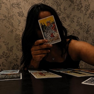 May include: A rectangular tarot card featuring a colorful illustration of a seated figure wearing a crown and robes, with the Cyrillic text ИМПЕРАТРИЦА printed at the bottom. The card is held upright, with several other tarot cards spread out on a dark surface.