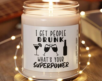 May include: A clear glass candle jar with a white label featuring black text that reads I GET PEOPLE DRUNK, WHAT'S YOUR SUPERPOWER, accompanied by icons of a martini glass, two clinking wine glasses, and a beer bottle, sitting on a wooden base.