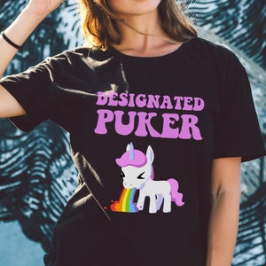 May include: Black t-shirt with a pink and white unicorn graphic and the text "Designated Puker" in pink letters.