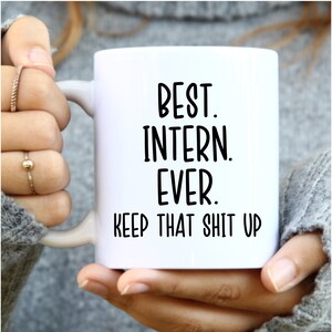 May include: White ceramic mug with the text "BEST. INTERN. EVER. KEEP THAT SHIT UP" in black, held by a person. The mug has a white handle and is a standard coffee mug size.