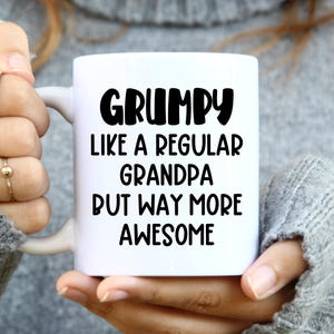 May include: A white ceramic mug featuring black text that reads: GRUMPY LIKE A REGULAR GRANDPA BUT WAY MORE AWESOME. The mug has a smooth, cylindrical body with a curved handle on the side, designed for holding hot beverages in a standard capacity.
