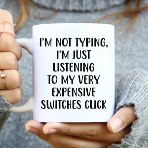 May include: White ceramic mug with the text "I'M NOT TYPING, I'M JUST LISTENING TO MY VERY EXPENSIVE SWITCHES CLICK" in black. The mug is held by a person wearing a grey jumper.