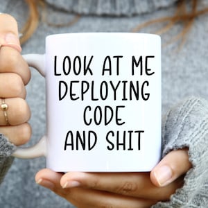 May include: White ceramic mug with the text "LOOK AT ME DEPLOYING CODE AND SHIT" in black. The mug is held by a person wearing a gray sweater. The mug has a white handle.