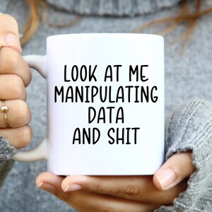 May include: White ceramic mug with the text "LOOK AT ME MANIPULATING DATA AND SHIT" in black, held by a person wearing a gray sweater. The mug has a standard handle and a glossy finish.