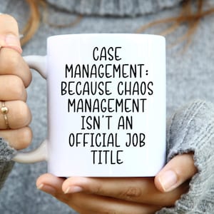 May include: White ceramic mug with the text "CASE MANAGEMENT: BECAUSE CHAOS MANAGEMENT ISN'T AN OFFICIAL JOB TITLE" in black. The mug is held by a person wearing a gray sweater and rings.