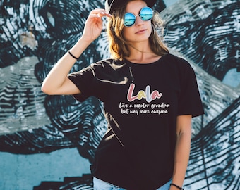 May include: A woman wearing a black t-shirt with the text "Lala Like a regular grandma but way more awesome" in colorful letters. She is wearing sunglasses and a black baseball cap.