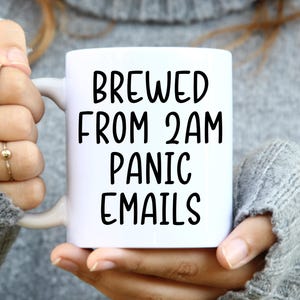 May include: White ceramic mug with the text "BREWED FROM 2AM PANIC EMAILS" in black. The mug is held by a person wearing a gray sweater. The mug has a white handle.