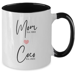 May include: White ceramic coffee mug with a black rim and handle. The mug has black text that reads "Mom Est. 1993" and "Coco Est. 2023" with a pink heart in between.