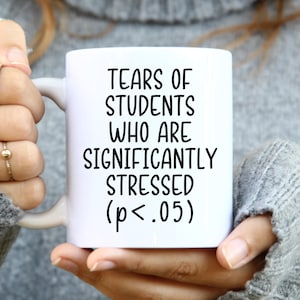 May include: A white ceramic mug featuring black printed text that reads: TEARS OF STUDENTS WHO ARE SIGNIFICANTLY STRESSED (p<.05). The mug has a standard handle and a smooth, glossy finish, displaying a humorous academic-themed design.
