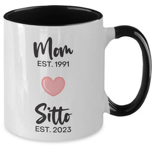 May include: White ceramic coffee mug with a black rim and handle. The mug has black text that reads "Mom EST. 1991" and "Sitto EST. 2023" with a pink heart in between.
