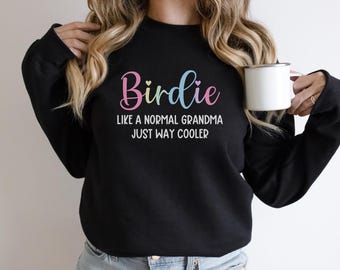 May include: Black sweatshirt featuring the word "Birdie" in pastel shades, with the text "Like a normal grandma just way cooler." A white mug is held. The sweatshirt is a casual style.