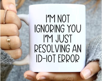 Könnte beinhalten: Ein weißer Keramikbecher mit dem schwarzen Aufdruck I'M NOT IGNORING YOU I'M JUST RESOLVING AN ID-IOT ERROR. Der Becher hat eine glatte Oberfläche und einen geschwungenen Henkel auf der linken Seite, konzipiert für heiße Getränke.