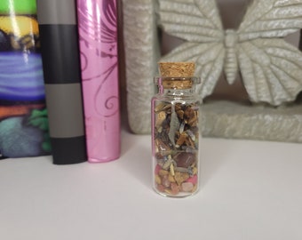 May include: A small glass bottle filled with various herbs, crystals, and stones. The bottle has a cork stopper and is sitting on a white surface.