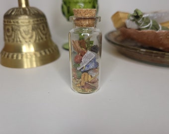 May include: A clear glass bottle filled with a variety of colorful dried herbs, crystals, and botanicals, sealed with a cork. The bottle is on a white surface, with a gold bell and a green glass nearby.