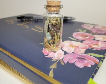 May include: A small glass bottle with a cork stopper filled with dried herbs, leaves, and a green stone. The bottle is sitting on a blue surface with pink flowers.