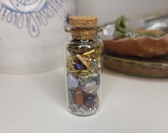 May include: A small glass bottle with a cork stopper filled with various crystals, dried herbs, and glitter. The bottle is clear and the contents are visible.