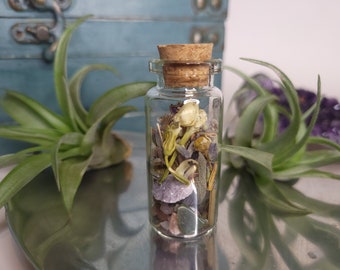 May include: A small glass bottle filled with dried flowers, herbs, and crystals. The bottle has a cork stopper and is sitting on a silver tray.