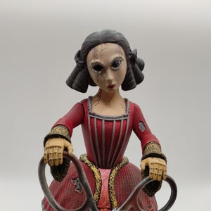 May include: A doll figurine with a cracked face and dark eyes, wearing a red dress and holding large silver scissors. The doll's dress has a layered skirt and gold accents.