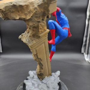 Pu&ograve; includere: Una action figure rossa e blu di Spider-Man che scala un pilastro di pietra rotto. La figura &egrave; in posa in una posa d'azione dinamica, con una gamba estesa e l'altra piegata. La figura &egrave; su uno sfondo grigio.