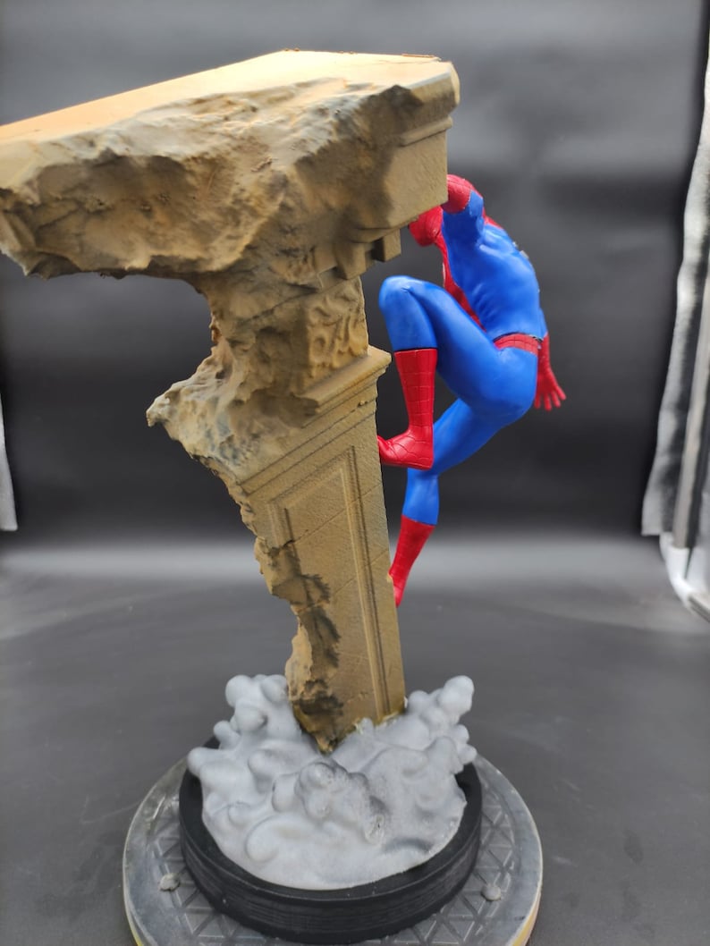 Pu&ograve; includere: Una action figure rossa e blu di Spider-Man che scala un pilastro di pietra rotto. La figura &egrave; in posa in una posa d'azione dinamica, con una gamba estesa e l'altra piegata. La figura &egrave; su uno sfondo grigio.