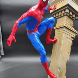 Pu&ograve; includere: Una action figure di Spider-Man, in rosso e blu, che scala un pilastro di pietra rotto. La figura &egrave; dettagliata con motivi a ragnatela e una maschera rossa.