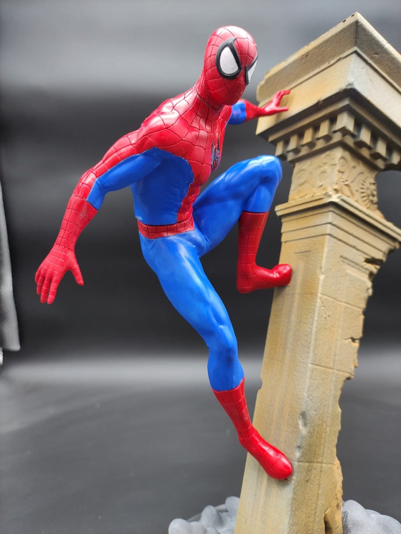 Pu&ograve; includere: Una action figure di Spider-Man, in rosso e blu, che scala un pilastro di pietra rotto. La figura &egrave; dettagliata con motivi a ragnatela e una maschera rossa.