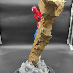 Pu&ograve; includere: Una action figure di Spider-Man rossa e blu &egrave; in posa mentre scala una formazione rocciosa rotta, grigia e marrone. La figura &egrave; in piedi su una base grigia e nera.